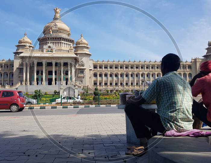 Bengaluru, Karnataka / India - August 20 2019: Two men sitting under a tree on footpath in front of the administrative building in Bangalore known as Vidhan Soudha during day time