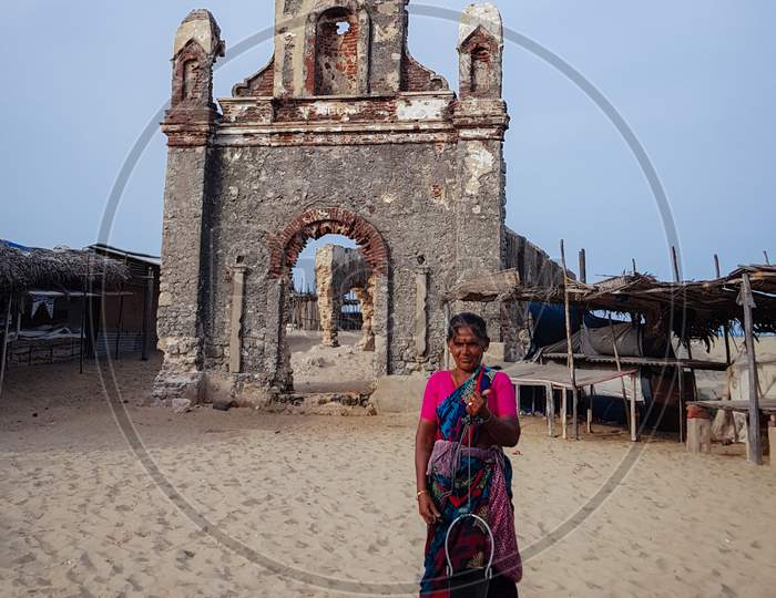 Dhanushkodi, India - 19 August 2018: An old woman drawing water from a well that gives potable water at a beach in Dhanushkodi. The church destroyed by the 1964 super cyclone is in the background.