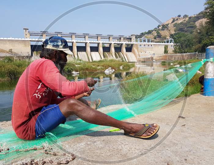 KRP Dam, Krishnagiri, Tamil Nadu, India - 25 Dec 2018: An unknown fisherman weaving a fishing net with the KRP Dam in the background