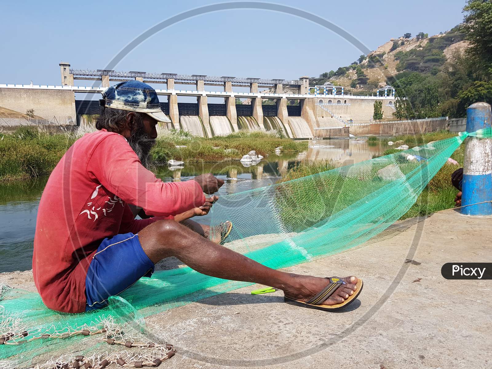 KRP Dam, Krishnagiri, Tamil Nadu, India - 25 Dec 2018: An unknown fisherman weaving a fishing net with the KRP Dam in the background