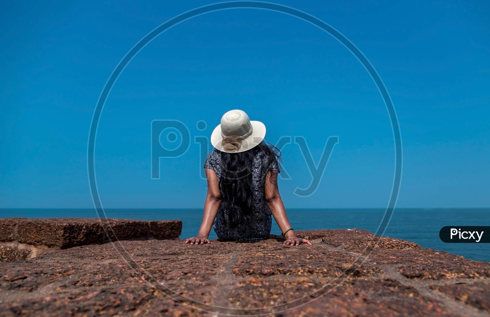 Beautiful wide angel view of a woman wearing a hat sitting on a wall looking at the sea on a sunny day with clear sky