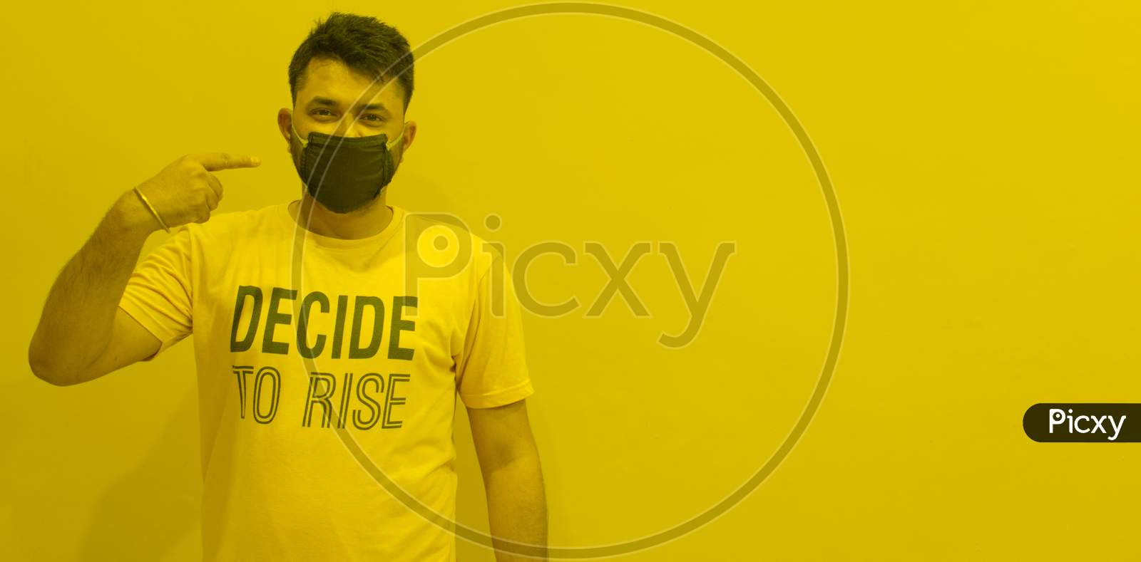 INDIAN MODEL WEARING BLACK MASK SHOWING FINGER TOWARDS PROTECTION MASK FROM CORONA WITH DECIDE TO RISE TSHIRT YELLOW BACKGROUND AND OPEN SPACE FOR TEXT