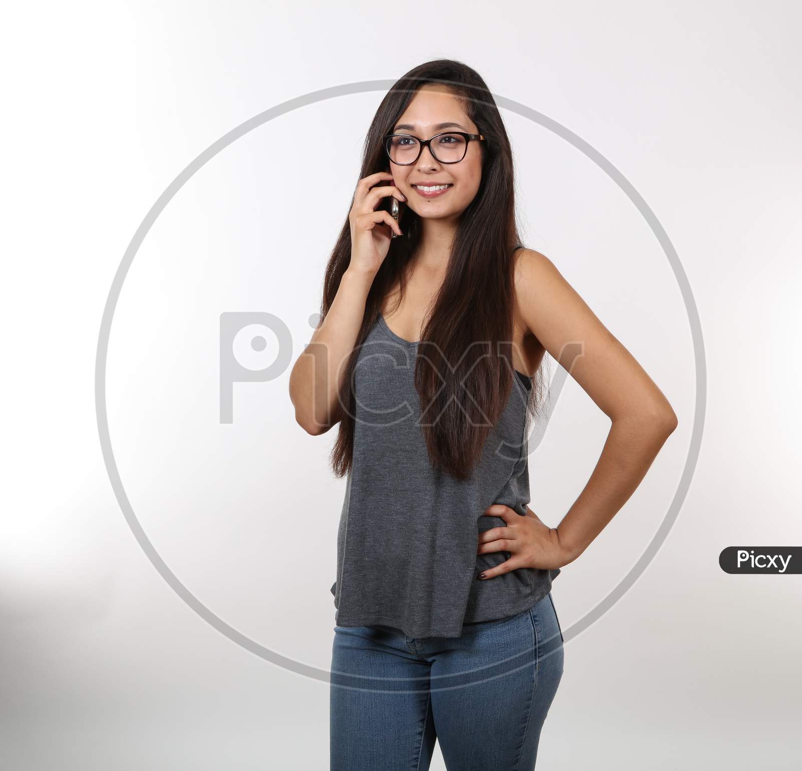 A Young Girl In Jeans And Glasses Smiles As She Hold Her Cell Phone Close To Her Ear.