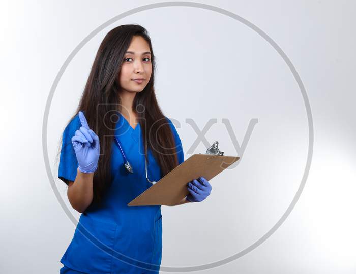 A Young Nurse In Blue Scrubs Holds A Clipboard And Points Her Finer Up.