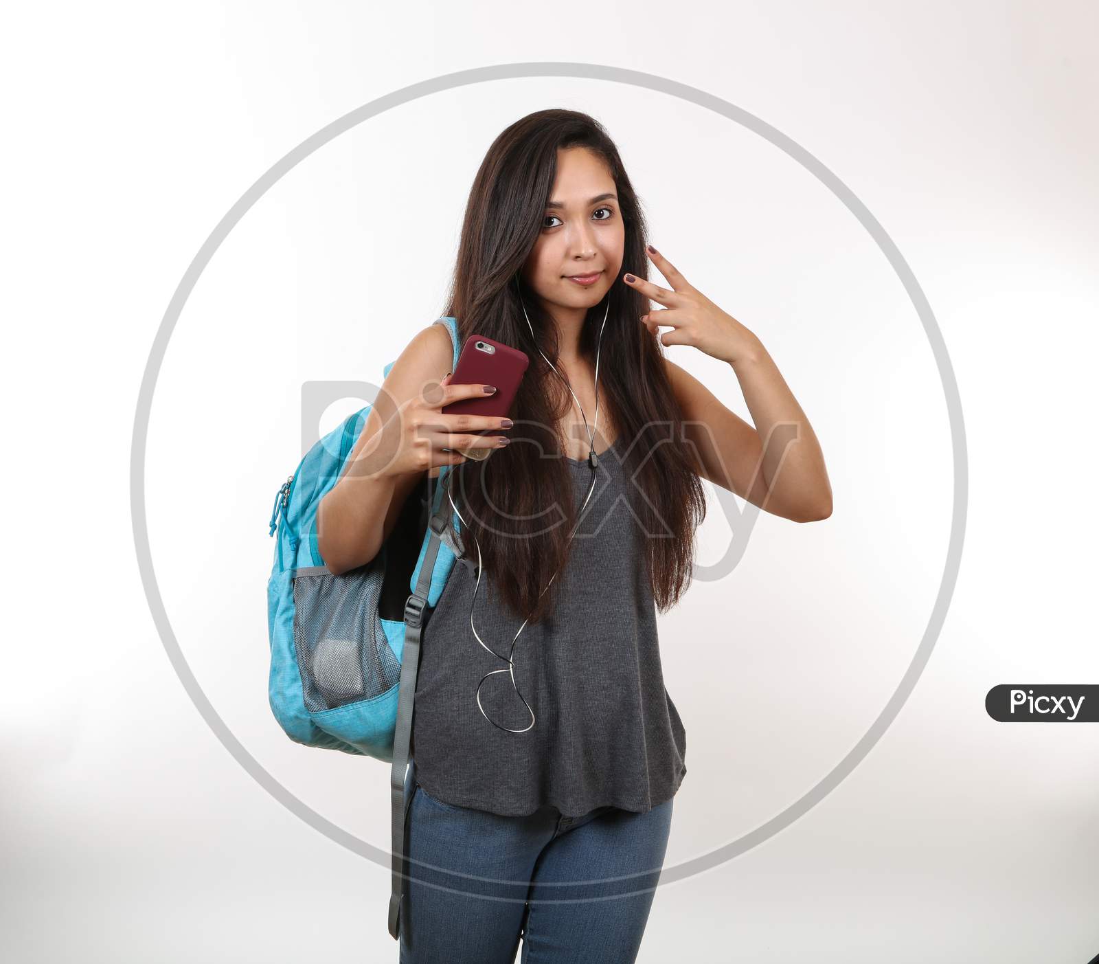 A Young Hispanic Student Wearing A Blue Backpack And Earphones, Holds Her Phone And Holds Up The Peace Sign With Her Hand.