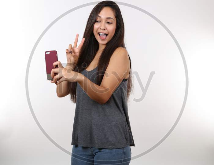 A Young Pretty Lady In Jeans And A Grey Shirt Holds The Peace Sign As She Takes A Picture On Her Cell Phone.