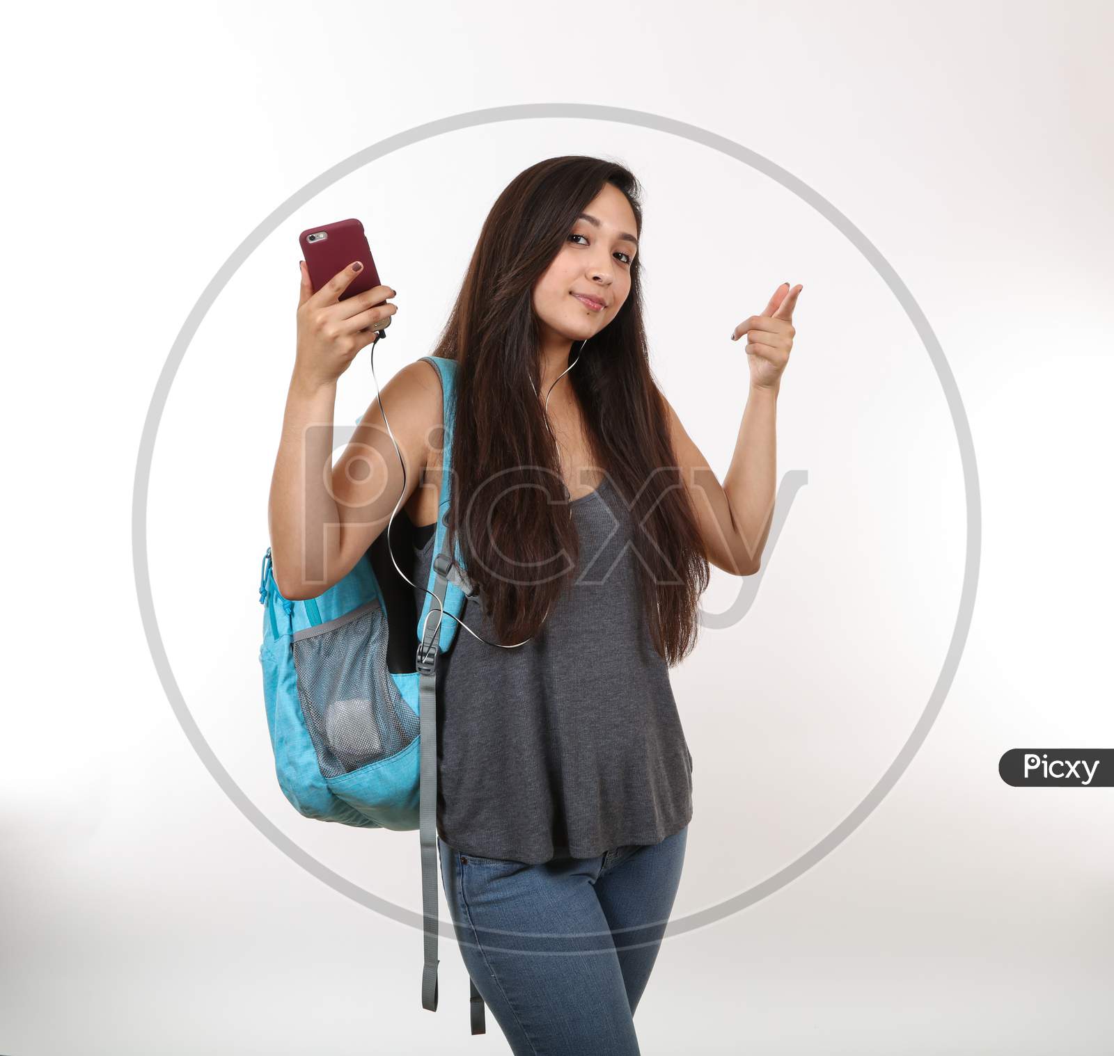 A Young Pretty Female Student Holds Her Phone As She Points Her Finger.  Student Is Also Wearing Earphones And A Backpack.