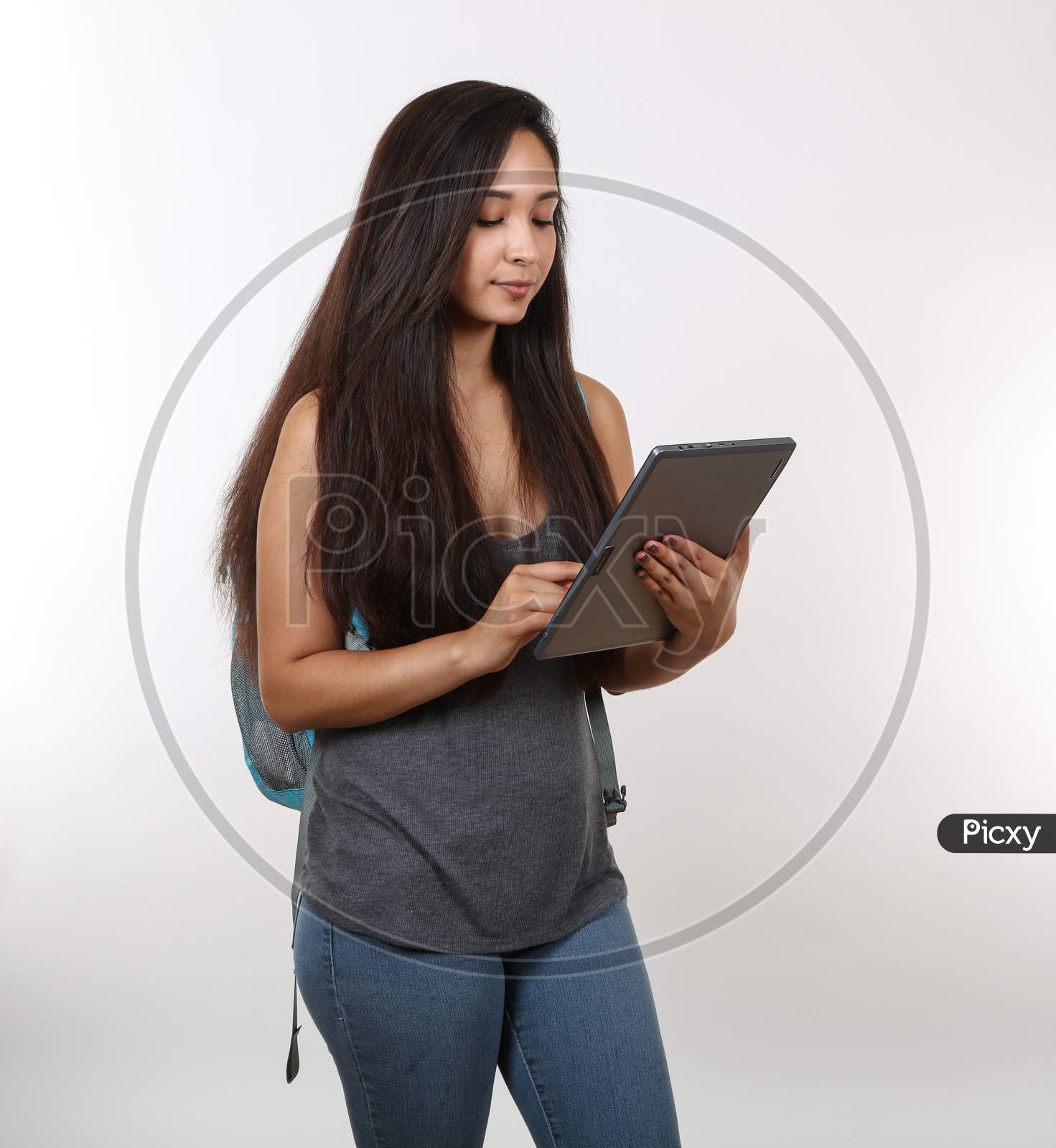 A Young Female Student In Jeans Views Her Tablet As She Holds Her Backpack.