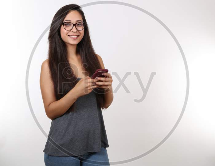 A Young Lady Wearing Glasses, Blue Jeans And A Grey Tank Top Holds Her Phone As She Smiles.