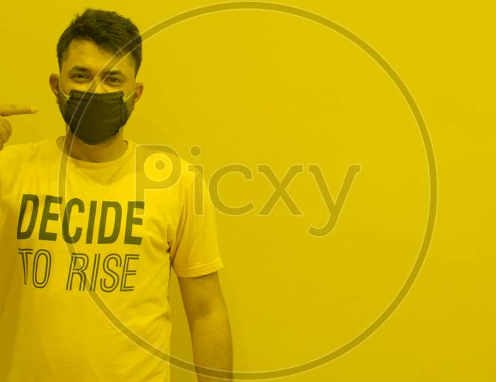 INDIAN MODEL WEARING BLACK MASK SHOWING FINGER TOWARDS PROTECTION MASK FROM CORONA WITH DECIDE TO RISE TSHIRT YELLOW BACKGROUND AND OPEN SPACE FOR TEXT
