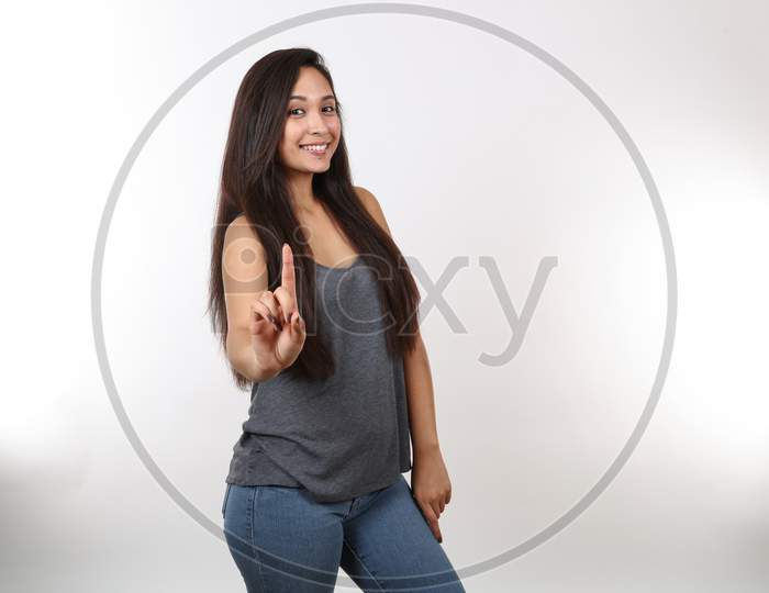 A Young Attractive Girl Smiles And Points Her Finger Up.  Wearing Blue Jeans And A Grey Tank Top.