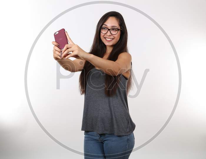 A Young Pretty Female Wearing Glasses Takes A Selfie With Her Cell Phone.