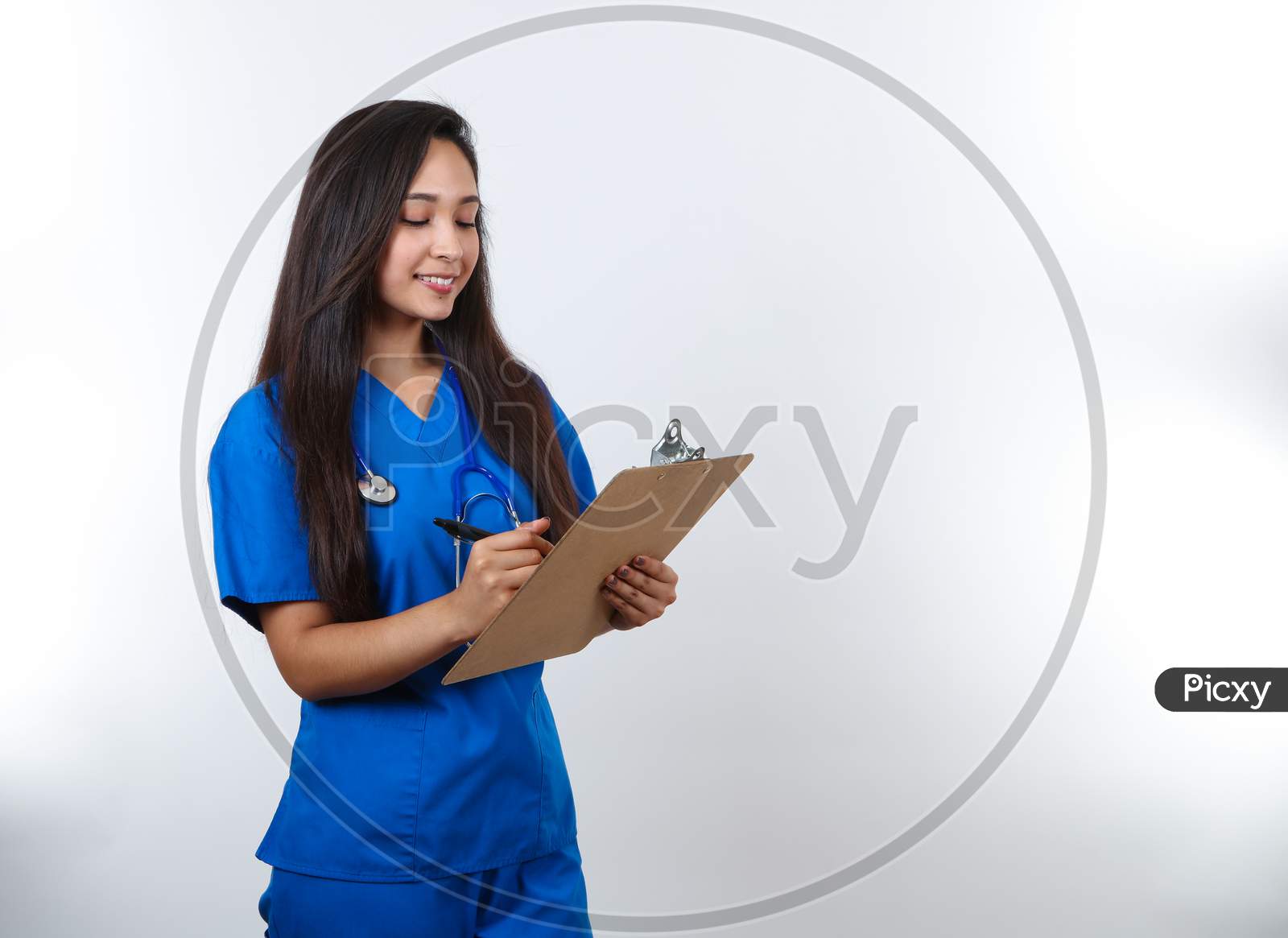 A Pretty Nurse In Blue Scrubs Writes Notes On Her Clip Board.