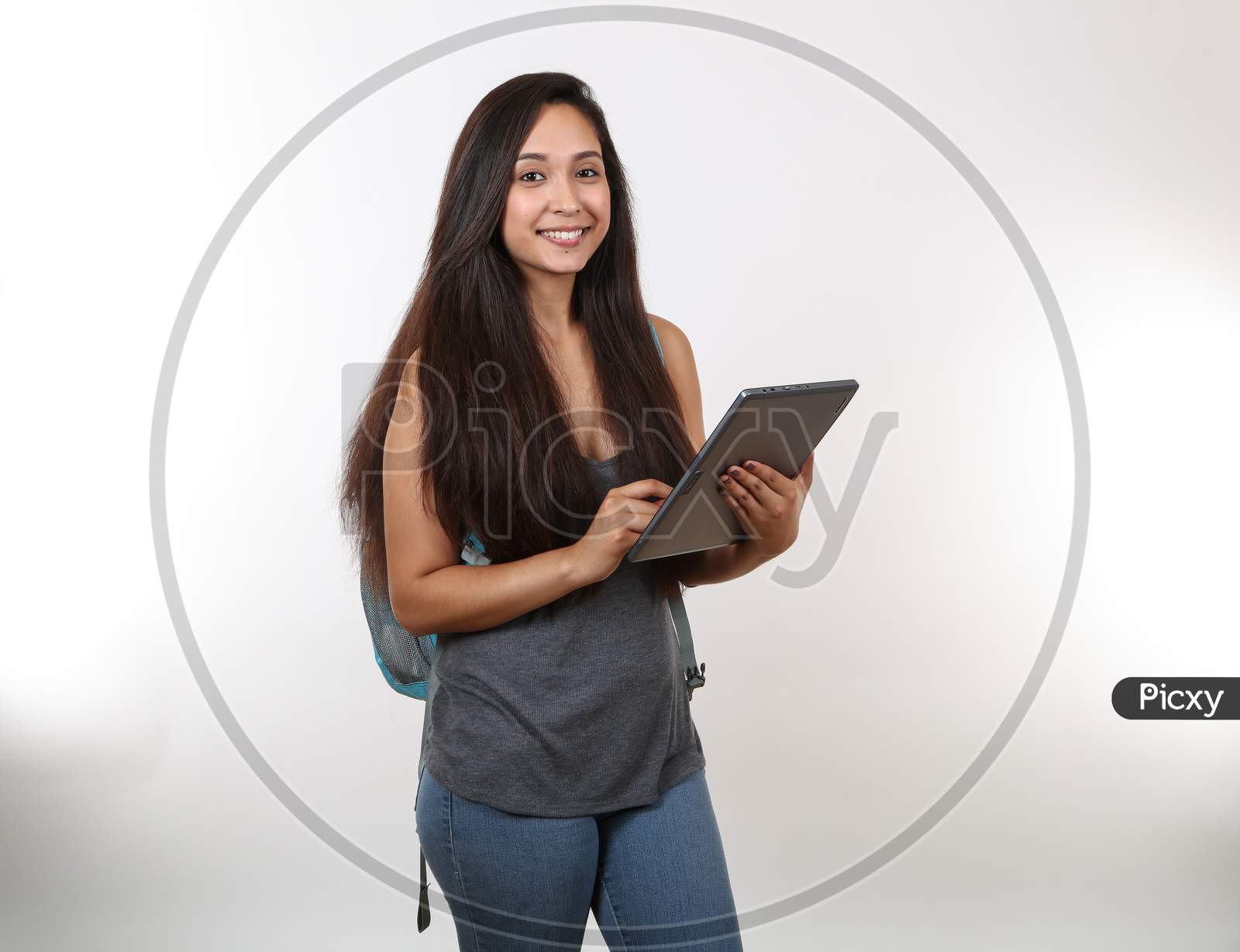 A Young Hispanic Student Wearing A Blue Backpack And Grey Shirt Smiles And She Holds Her Digital Device.