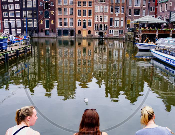 Tourists Taking A Break In Amsterdam City Center, With Iconic Colorful Amsterdam Buildings And Canals In The Background