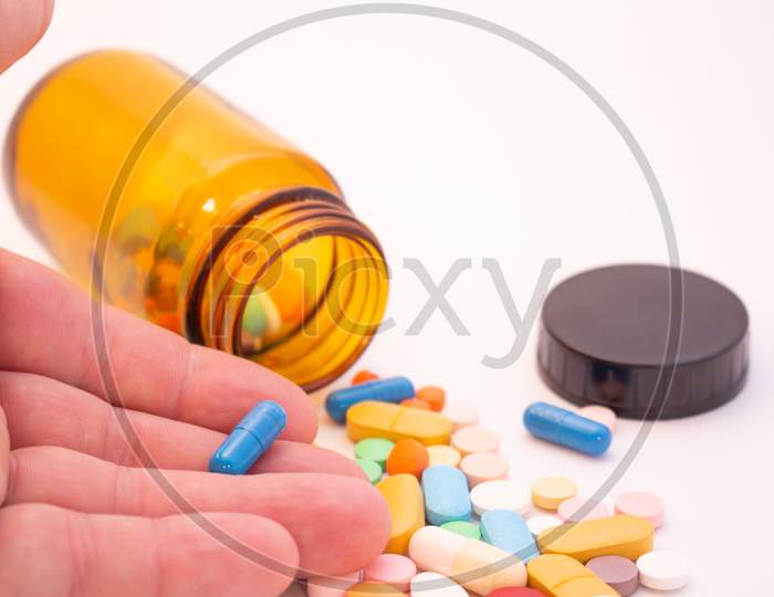 Medications In The Form Of Pills And Capsules On A White Background. Brown Glass Vessel. Hand Of A Man Holding A Blue Capsule.