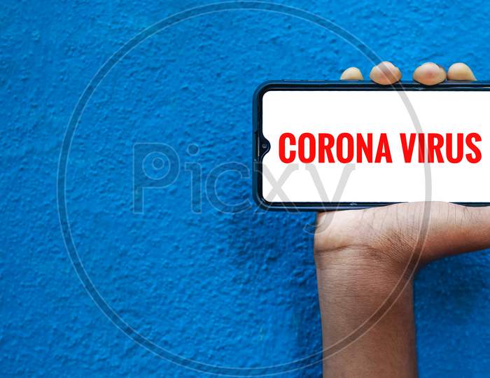 Corona Virus / Covid-19 Wording On Smart Phone Screen Isolated On Blue Background With Copy Space For Text. Person Holding Mobile On His Hand And Showing Front Of The Screen For Corona Virus Wording.