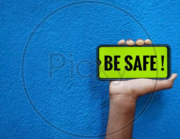 Be Safe World On Smart Phone Screen Isolated On Blue Background With Copy Space For Text. Person Holding Mobile On His Hand And Showing Front Of Be Safe Corona Virus.