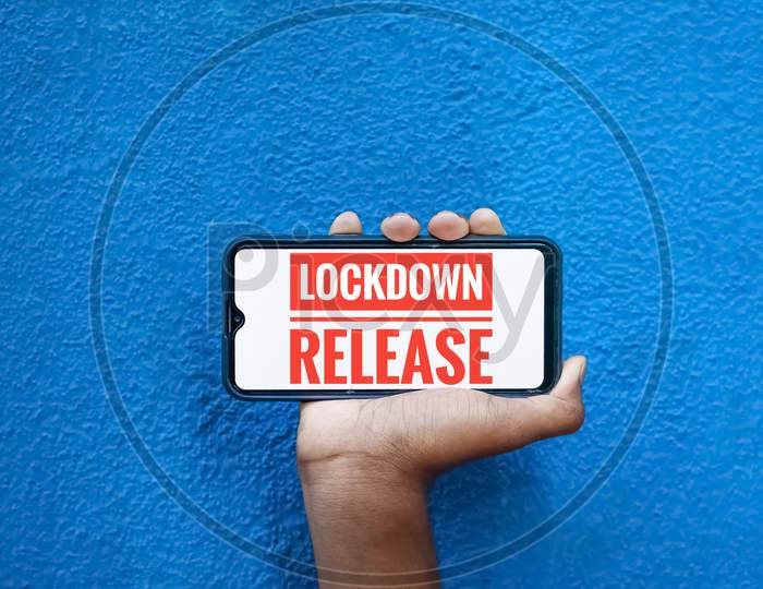 Lock Down Release Wording On Smart Phone Screen Isolated On Blue Background With Copy Space For Text. Person Holding Mobile On His Hand And Showing Front Screen. Lock Down Release For Corona Virus .