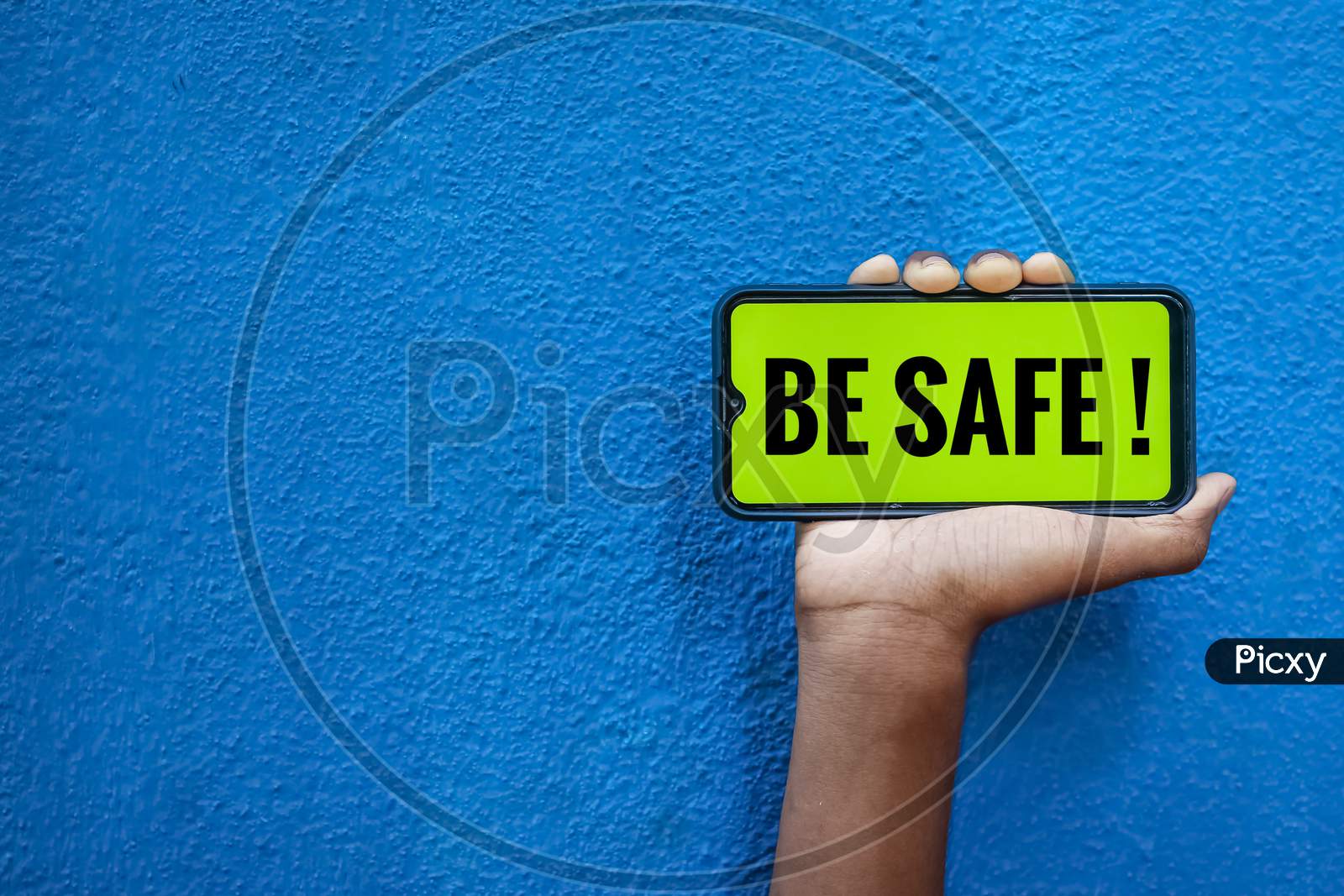 Be Safe World On Smart Phone Screen Isolated On Blue Background With Copy Space For Text. Person Holding Mobile On His Hand And Showing Front Of Be Safe Corona Virus.