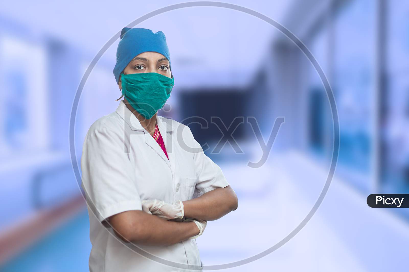 Portrait Of Female Medical Worker Doctor Wearing Surgical Mask And Cap Standing Crossed Arms Outside Hospital Corridor. Covid-19, Coronavirus Pandemic.