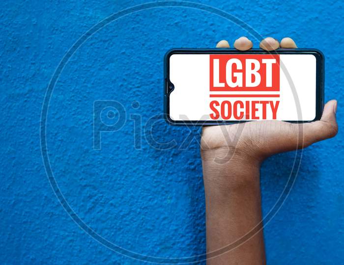 Lgbt Society Word On Smart Phone Screen Isolated On Blue Background With Copy Space For Text. Person Holding Mobile On His Hand And Showing Front Of Lgbt Society.(Lesbi, Gay, Bisexual, Transgender).