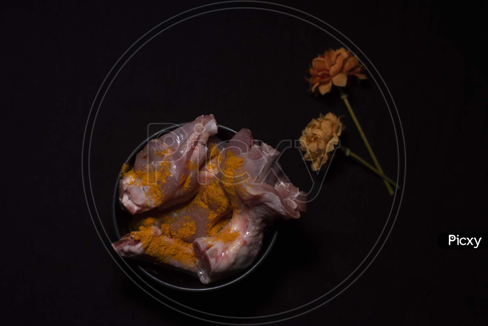 Top Down Image Of A Bowl Of Raw Chicken Pieces Sprinkled With Turmeric Powder Decorated With Dried Flowers In A Dark Copy Space Background. Food And Product Photography.