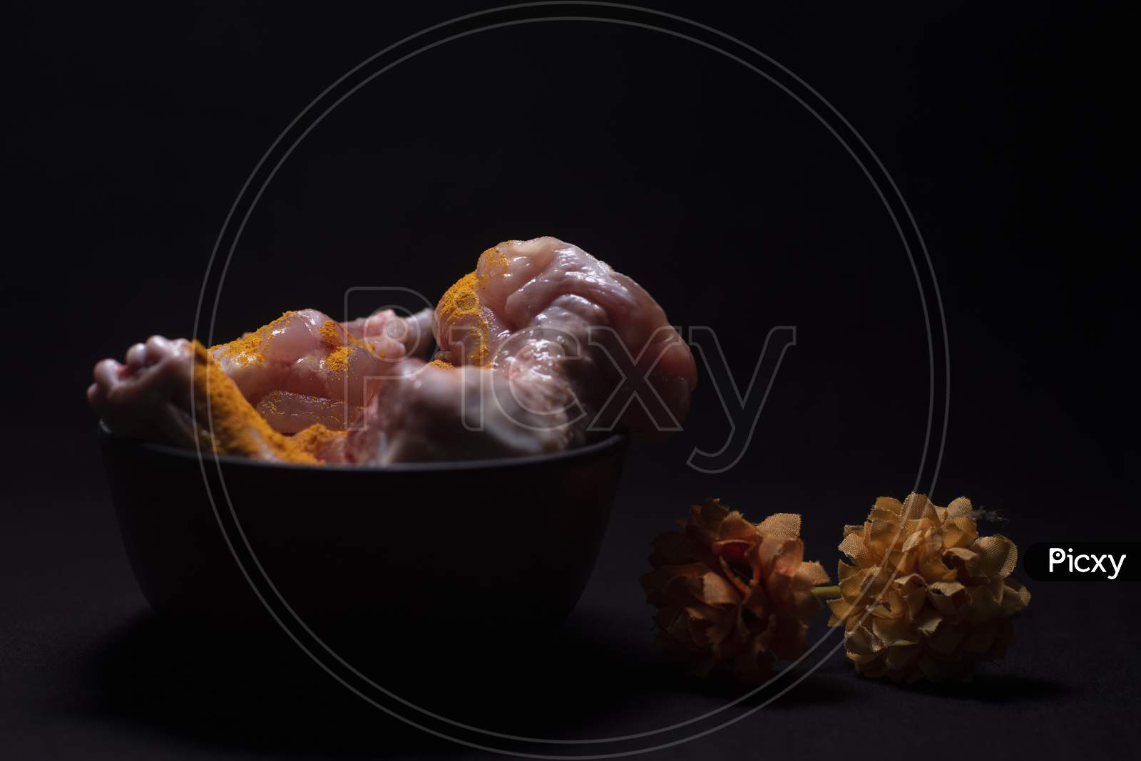 A Bowl Of Raw Chicken Pieces Sprinkled With Turmeric Powder Decorated With Dried Flowers In A Dark Copy Space Background. Food And Product Photography.