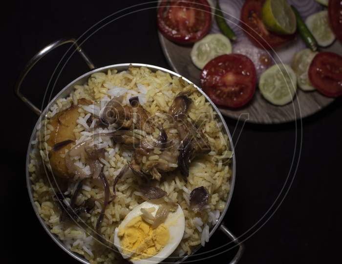 Top Down Image Of Indian Spicy Dish Biryani With Egg, Potato And Chicken In A Pot Along With Fresh Salads In Dark Copy Space Background. Food And Object Photography.