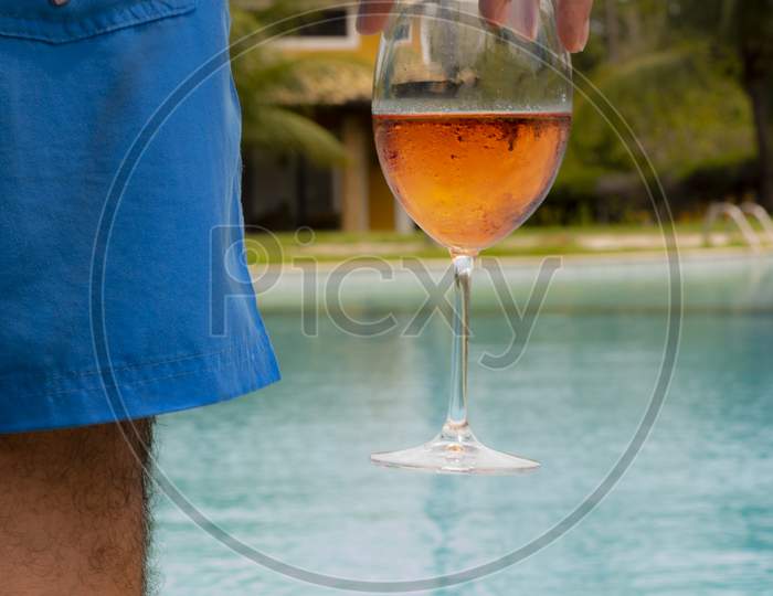 Man Holding A Glass Goblet With Fresh Rose Wine Next To A Huge Pool Of Crystal Clear Water. Luxury Wealth And Vacation Concept.