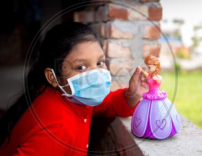 Bangladesh – April 16, 2020: Asian Girl Is Wearing A Surgical Face Mask And Playing With A Pink Barbie Toy On Their Own Home At Dhaka.