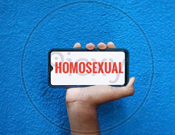 Homosexual Word On Smart Phone Screen Isolated On Blue Background With Copy Space For Text. Person Holding Mobile On His Hand And Showing Front Of Homosexual.(Lesbi, Gay, Bisexual, Transgender).