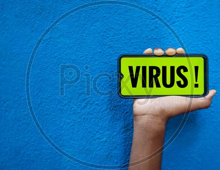 Virus / Covid-19 Wording On Smart Phone Screen Isolated On Blue Background With Copy Space For Text. Person Holding Mobile On His Hand And Showing Front Of The Screen For Corona Virus Wording.