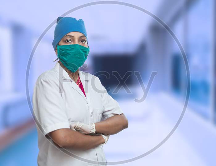 Portrait Of Female Medical Worker Doctor Wearing Surgical Mask And Cap Standing Crossed Arms Outside Hospital Corridor. Covid-19, Coronavirus Pandemic.
