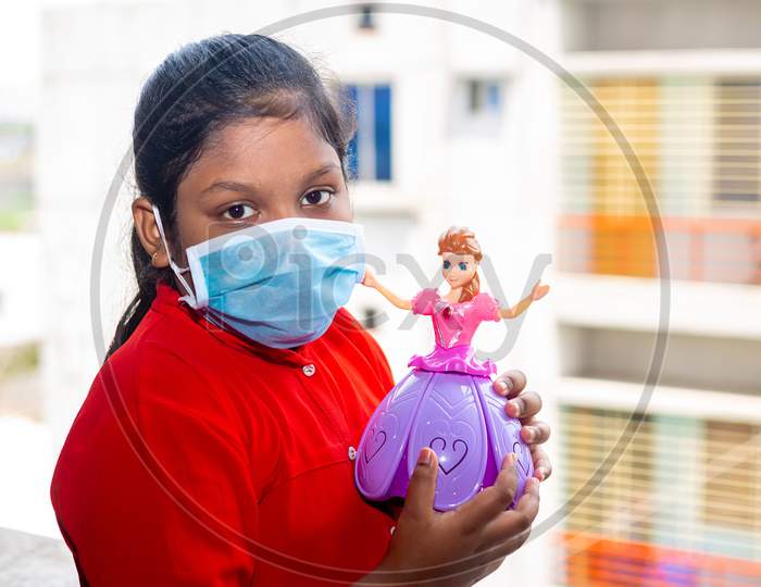 Bangladesh – April 16, 2020: Asian Girl Is Wearing A Surgical Face Mask And Playing With A Pink Barbie Toy On Their Own Home At Dhaka.