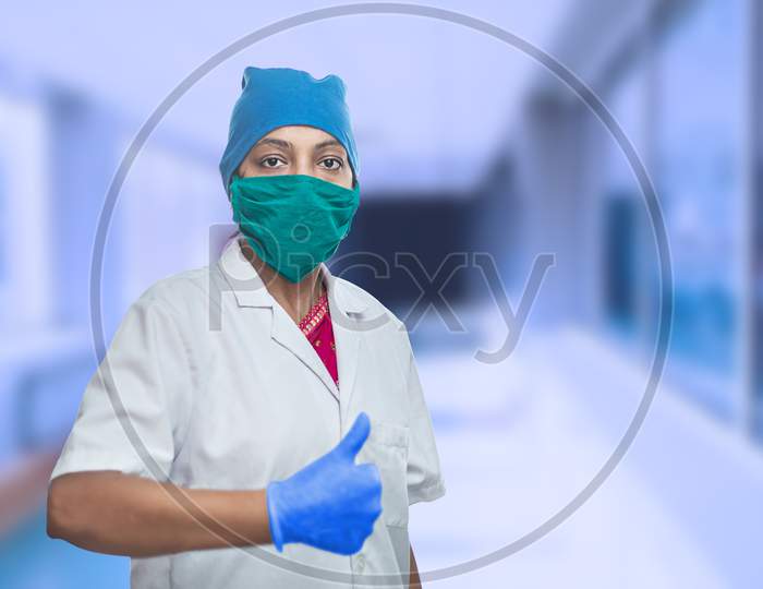 Portrait Of Female Medical Worker Doctor Wearing Surgical Mask And Cap Doing Thumbs Up, Ok Sign, Excellent Sign. Outside Hospital Corridor. Covid-19, Coronavirus Pandemic.