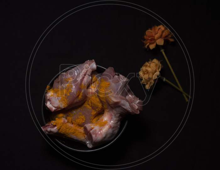 Top Down Image Of A Bowl Of Raw Chicken Pieces Sprinkled With Turmeric Powder Decorated With Dried Flowers In A Dark Copy Space Background. Food And Product Photography.
