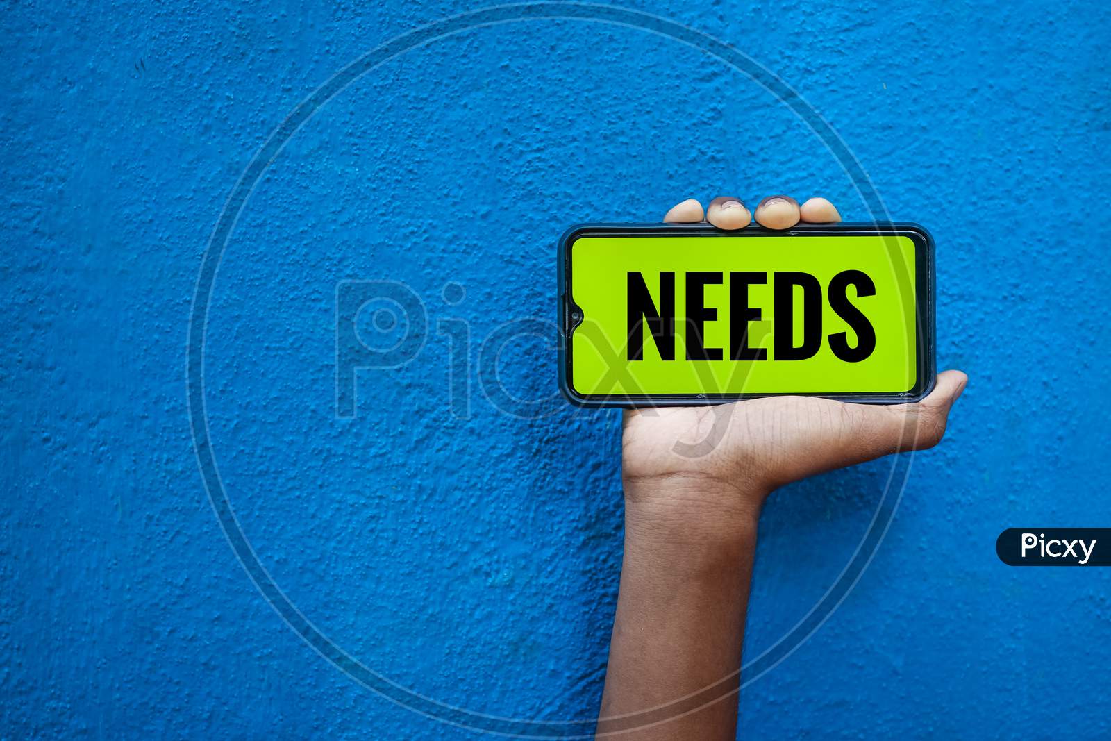 Needs Wording On Smart Phone Screen Isolated On Blue Background With Copy Space For Text. Person Holding Mobile On His Hand And Showing Front Of The Screen Word Needs.