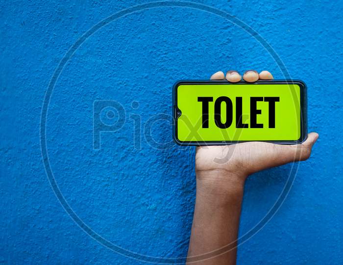 Tolet Wording On Smart Phone Screen Isolated On Blue Background With Copy Space For Text. Person Holding Mobile On His Hand And Showing Front Of The Screen Word Tolet Rental House.