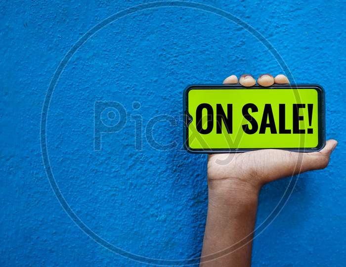 On Sale Wording On Smart Phone Screen Isolated On Blue Background With Copy Space For Text. Person Holding Mobile On His Hand And Showing Front Of The Screen Word On Sale