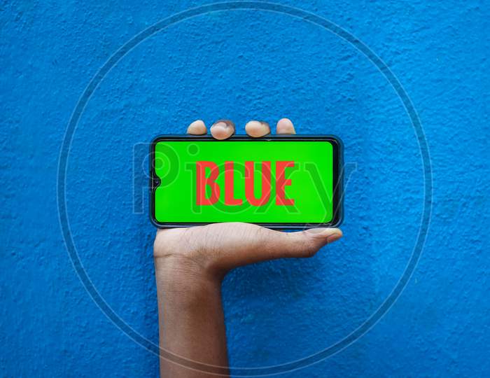 Blue Word On Orange Color In Smart Phone Screen Isolated On Blue Background With Copy Space For Text. Person Holding Mobile On His Hand And Showing Front Of The Screen Word Blue.