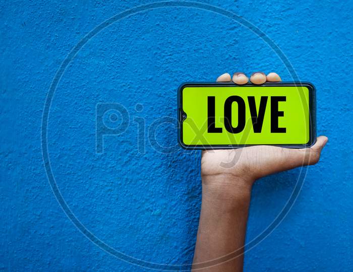 Love Wording On Smart Phone Screen Isolated On Blue Background With Copy Space For Text. Person Holding Mobile On His Hand And Showing Front Of The Screen Word Love.