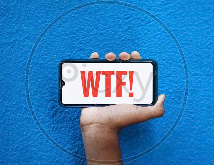 Wtf Wording On Smart Phone Screen Isolated On Blue Background With Copy Space For Text. Person Holding Mobile On His Hand And Showing Front Of The Screen Concept Wtf Scold.