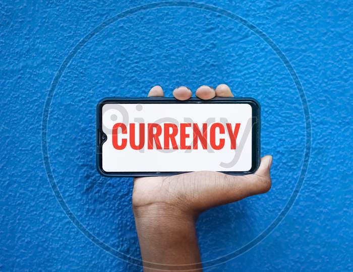 Currency Wording On Smart Phone Screen Isolated On Blue Background With Copy Space For Text. Person Holding Mobile On His Hand And Showing Front Of The Screen Concept Money Currency