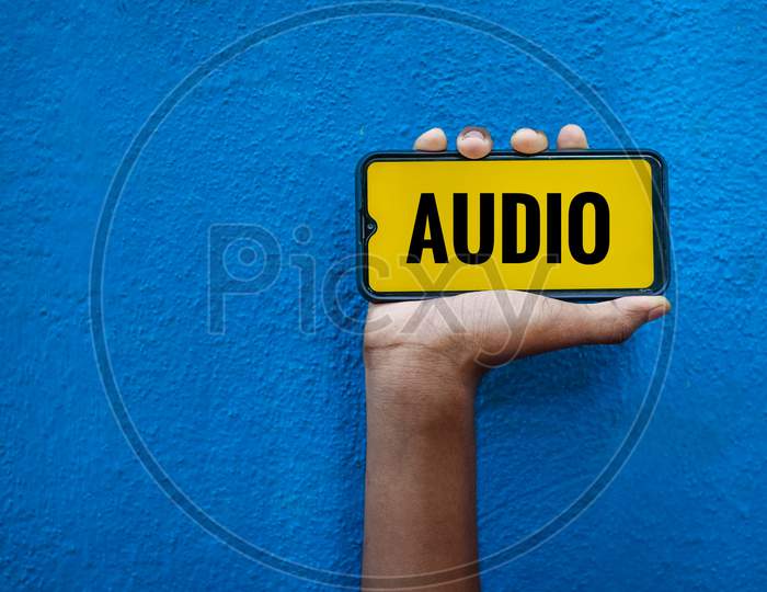 Audio Word On Smart Phone Screen Isolated On Blue Background With Copy Space For Text. Person Holding Mobile On His Hand And Showing Front Of The Screen Wording Audio.