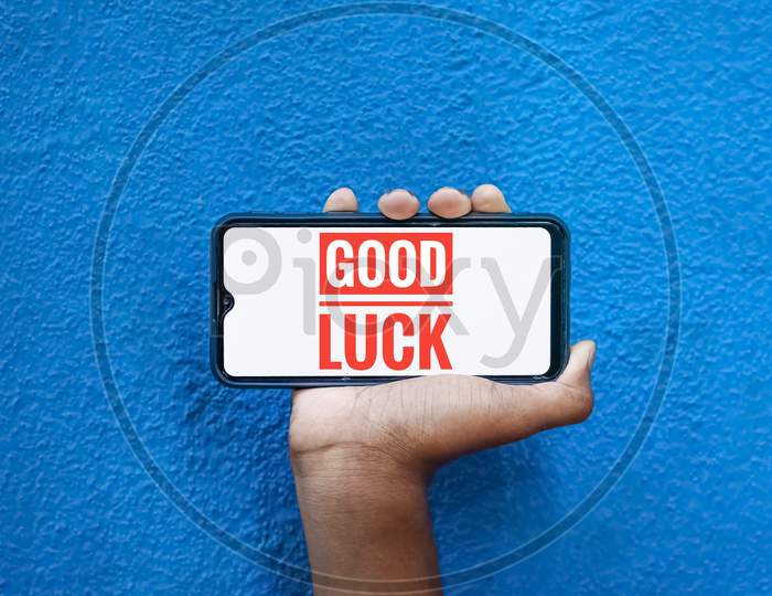 Good Luck Wording On Smart Phone Screen Isolated On Blue Background With Copy Space For Text. Person Holding Mobile On His Hand And Showing Front Of The Screen Word Good Luck