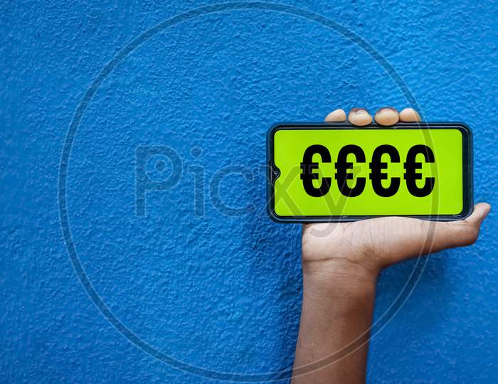 Eur Currency Symbol On Smart Phone Screen Isolated On Blue Background With Copy Space For Text. Person Holding Mobile On His Hand And Showing Front Of The Screen Eur Currency Symbol.