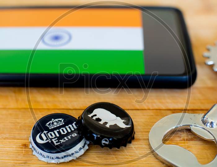 Mobile Phone With India Flag And Two Beer Bottle Caps And An Opener On Wooden Board