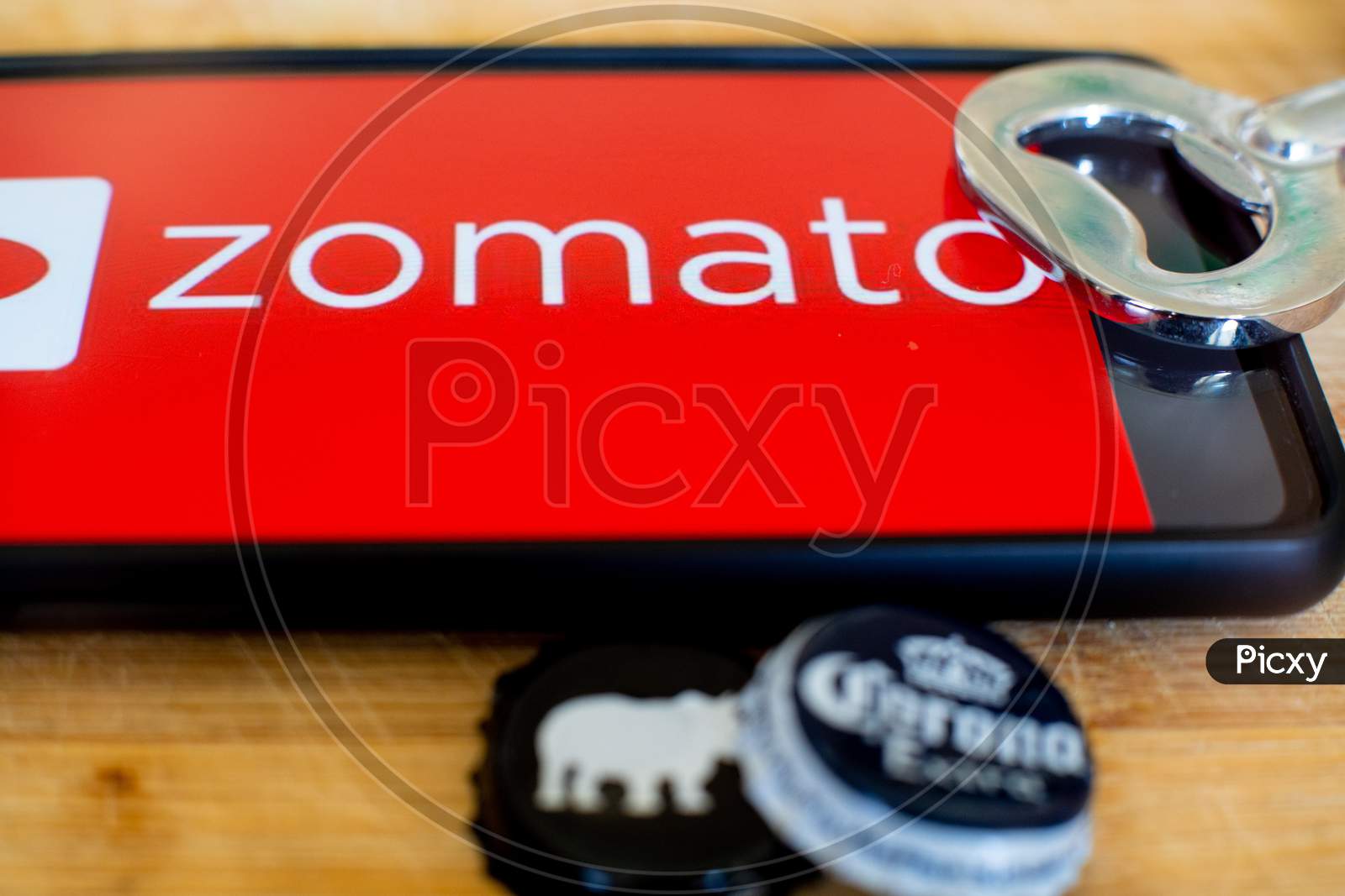 Mobile Phone Showing Zomato On A Wooden Platform With Beer Bottle Caps And An Opener On The Side