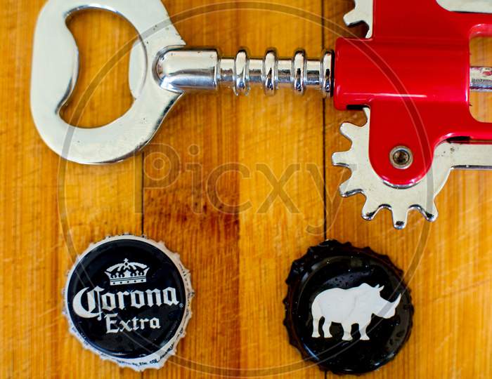 Shot Of Wooden Board With Mobile Phone, Bottle Opener, And Beer Bottle Caps Of Corona And White Rhino